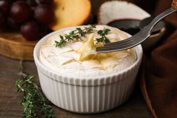 Eating tasty baked camembert with fork from bowl at wooden table, closeup