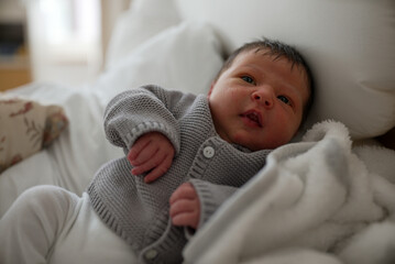 Awake newborn baby with dark hair in a gray knitted sweater, lying on a white blanket. The baby is...