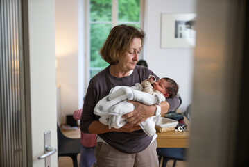 Grandmother holding a newborn baby wrapped in a white blanket in a cozy home setting. The grandmother is smiling warmly at the baby, creating a moment of generational bonding and love.