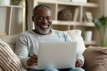 A happy African American middle-aged man is using a laptop while sitting on the sofa at home. The mature older user looks at the computer screen, browsing the internet and doing ecommerce shopping on