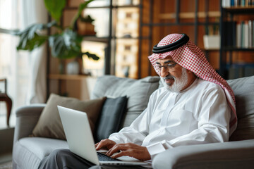 A happy Arabian middle-aged man is using a laptop while sitting on the sofa at home. The mature older user looks at the computer screen, browsing the internet and making ecommerce purchases on a