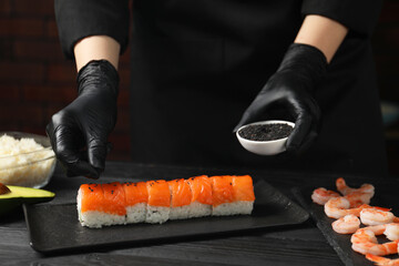 Chef in gloves adding sesame seeds onto tasty sushi rolls at black wooden table, closeup