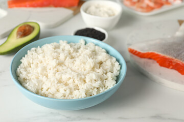 Cooked rice in bowl and other ingredients for sushi on white marble table, closeup