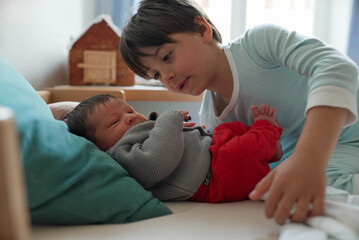 Older sibling leaning over newborn baby, both engaged in a curious and tender interaction. The room...