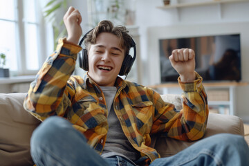 A joyful and spirited Caucasian teen guy, representative of Gen Z hipster culture, wears headphones as he dances at home. He's immersed in the music playing on his mobile phone, feeling funky and