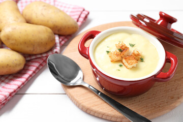 Tasty potato soup with croutons in ceramic pot and spoon on white wooden table