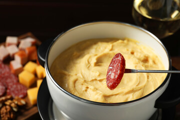 Dipping piece of sausage into fondue pot with melted cheese at table, closeup