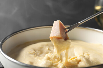 Dipping piece of ham into fondue pot with melted cheese on grey background, closeup