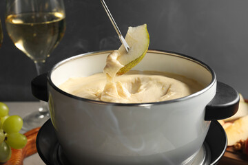 Dipping piece of pear into fondue pot with melted cheese at table on grey background, closeup