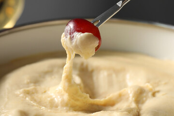 Dipping piece of grape into fondue pot with melted cheese on grey background, closeup