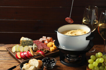 Dipping piece of sausage into fondue pot with melted cheese at wooden table with products, closeup