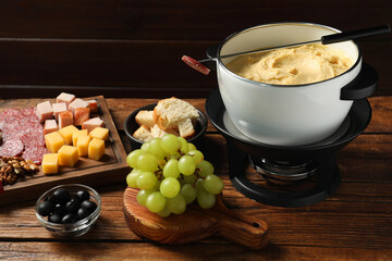 Fork with piece of sausage, melted cheese in fondue pot and other products on wooden table