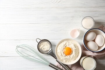 Flour with yolk in bowl and other ingredients for dough on white wooden table, flat lay. Space for text