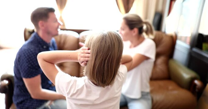 Teen girl covers ears with hands to avoid listening to argument of parents. Man and woman sit on sofa quarreling in presence of daughter slow motion