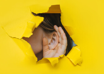 Female left ear and hand close-up. Copy space. Torn paper, yellow background. The concept of eavesdropping, espionage, gossip and the yellow press.