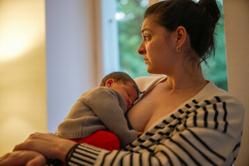 Mother holding her sleeping newborn baby close to her chest, looking contemplatively out of a...