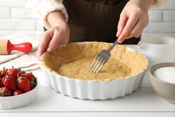 Shortcrust pastry. Woman making holes in raw dough with fork at white wooden table, closeup