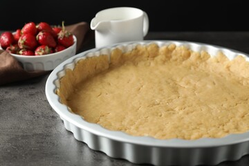Making shortcrust pastry. Raw dough in baking dish, strawberries and milk on grey table, closeup