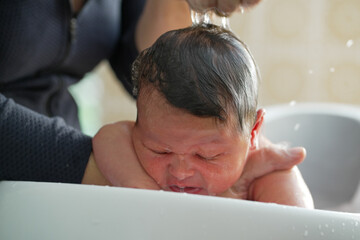 A baby leans forward in a white basin while getting a gentle hair wash from a parent. The baby’s...