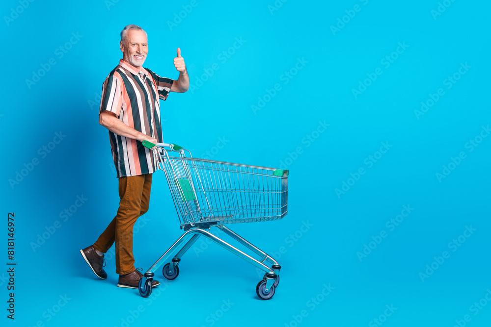 Poster photo of nice senior man wear summer striped clothes walk pushcart thumb up empty space isolated on 