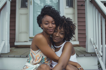 Fototapeta premium Two Black sisters sitting on stairs in front of a house, sharing a heartfelt embrace. Capturing the essence of sisterly love and the strong relationship between siblings.