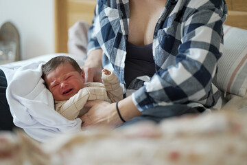 Mother in plaid shirt sits on bed, cradling her crying baby in beige clothes. The mother’s...