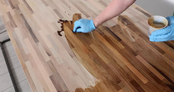 Worker in gloves polishes with varnish wooden floor surface standing at workplace in premise. Man uses liquid lacquer for processing wood slow motion
