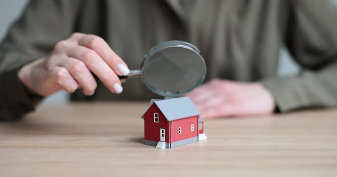 Woman holds magnifier glass above model of townhouse sitting at table in light premise. Inspection of property building slow motion. Insurance concept