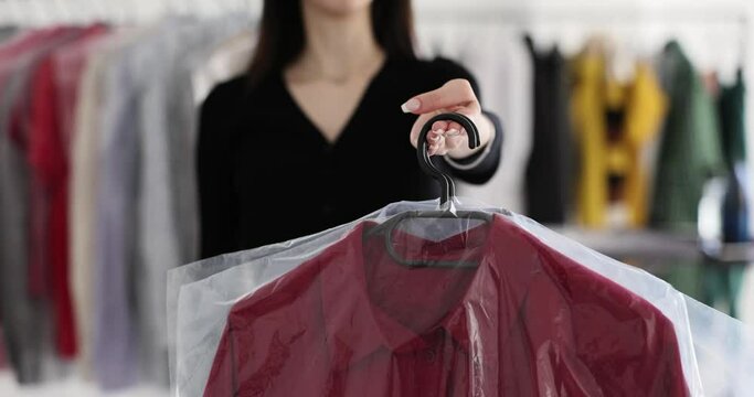 Female manager of dry cleaning holds red men shirt standing in company office. Brunette employee offers client to pick up clean clothes slow motion