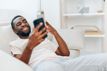 Happy African American man resting on a black sofa, using his mobile phone for communication and...