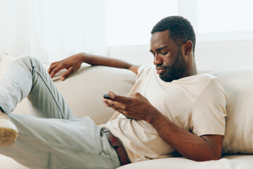 Happy African American Man Typing on His Black Smartphone While Relaxing on a Modern Sofa in His...