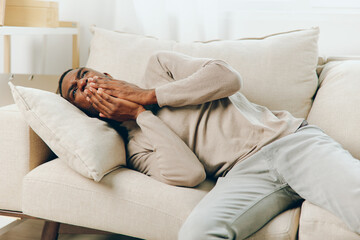 Obraz premium Stressed African American Man with Headache Sitting on Couch at Home The man, exhausted and sicklooking, clutches his head in pain as he rests on the sofa in his living room The combination of