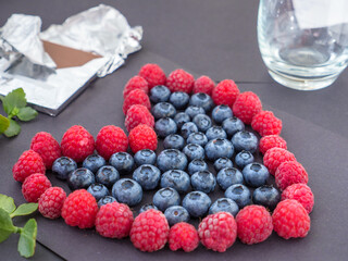 Heart shape love sign made by fresh raspberries and blueberries on black paper. Chocolate bar in foil, leaves and a glass in the background. Love and Valentine's day concept.