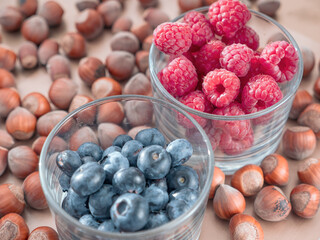 Closeup of fresh ripe raspberries and blueberries in low transparent glasses and closed hazelnuts at the blurry background.