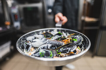 A close-up of a pan of cooked mussels garnished with herbs, held by a person in a kitchen setting