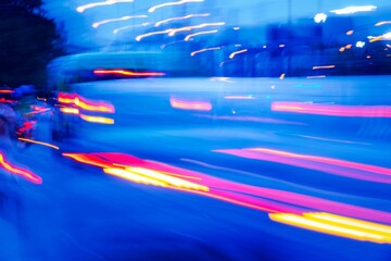 a busy street with cars and a bus. The image has a sense of motion and energy, as if it were captured in a time-lapse. The blue and red colors of the cars