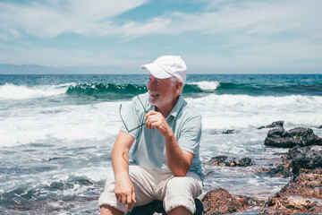 Carefree senior relaxed man barefoot wearing sunglasses and cap sitting on a rocky beach admiring...