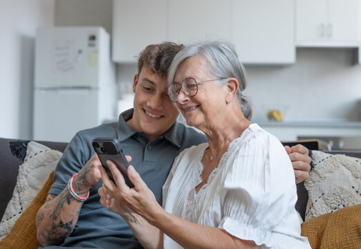 Happy grandmother learning how to use mobile phone technology from young grandson, handsome smiling boy teaching grandma shop online, book travel and vacation