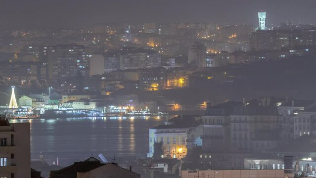 Aerial view of Lisboa downtown night timelapse. Panoramic of Baixa, Rossio and Chiado rooftops from above. Illuminated buildings. Almada on the other side of Tejo river. Portugal