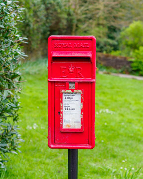 Red royal mail postal box front view with green grass behind in a English village location