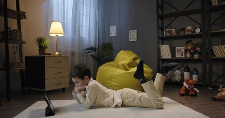 Little kid boy use digital tablet in the evening, watch streaming service, enjoy video content in social media. Smiling boy relax lay on the floor with tablet device - Powered by Adobe