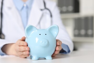 Doctor with piggy bank at white table indoors, closeup. Space for text