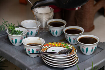 A jebena sits on a grass-covered table with small, handle-less cups arranged around it. Roasted coffee beans, a mortar and pestle, and incense smoke create a traditional Ethiopian coffee ceremony