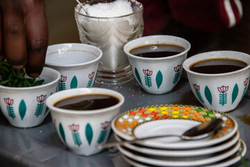 A jebena sits on a grass-covered table with small, handle-less cups arranged around it. Roasted coffee beans, a mortar and pestle, and incense smoke create a traditional Ethiopian coffee ceremony