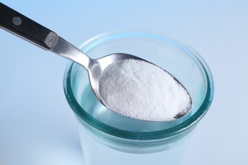 Spoon with baking soda over glass of water on light blue background, closeup