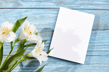 A single white blank card positioned next to white Alstroemeria flowers on a blue painted wooden...