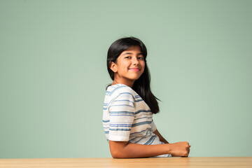 Indian asian small girl sitting at table against plain green background