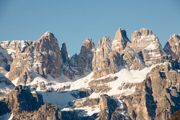 The stunning mountain Paganella in Italy, with rugged, jagged peaks partially covered in snow. Steep rock faces and snow-dusted ridges. Majestic landscape under a clear blue sky.
