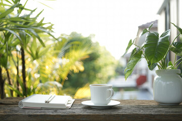 White coffee cup on wooden tray and notebook and Philodendron plant on wooden table under sunlight