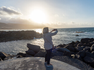 Mature woman taking photo with her cell phone by the sea at sunset